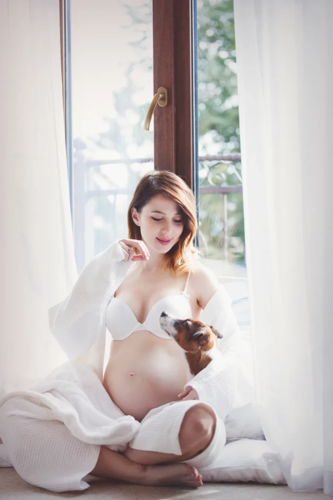 A pregnant woman posing near a window for her maternity boudoir photo session.